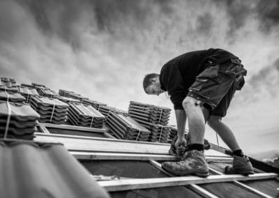 Portrait en action d'un artisan couvreur qui découpe des voliges sur un toit. Une photo originale en noir et blanc, le ciel est chargé.
