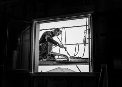 Zingueur en train de souder un chêneau. La photo de l'artisan est en noir et blanc, l'image originale montre la scene a travers une fenêtre cerclée d'un fond noir